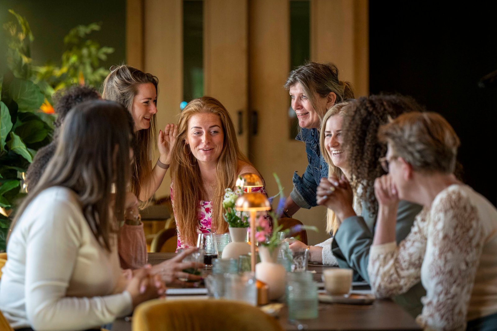 A group of joyful women engaging in conversation around a dining table in a warm, well-lit room.